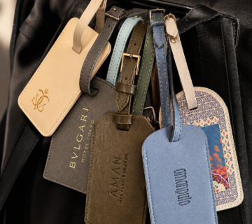 A collection of luxury hotel luggage tags hanging from a black travel bag, including tags from Aman, Bulgari and other high end hotel brands 