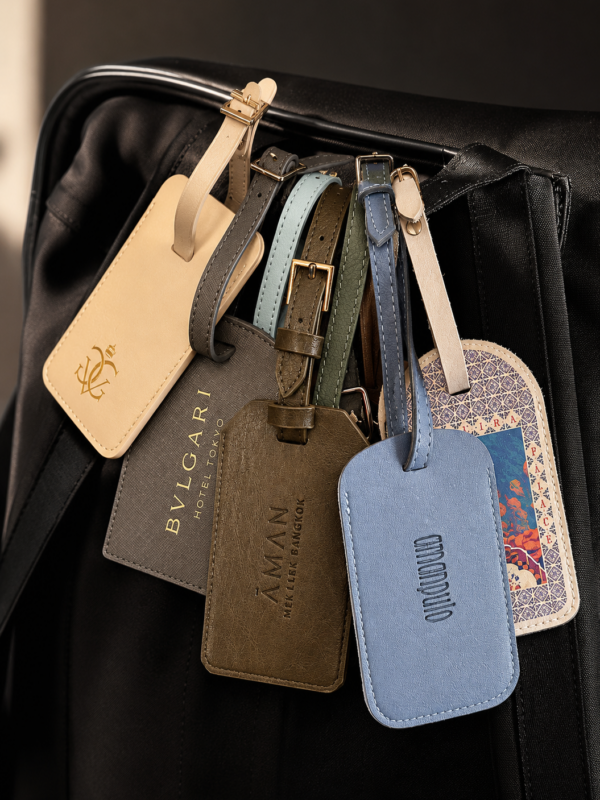 A collection of luxury hotel luggage tags hanging from a black travel bag, including tags from Aman, Bulgari and other high-end hotel brands.